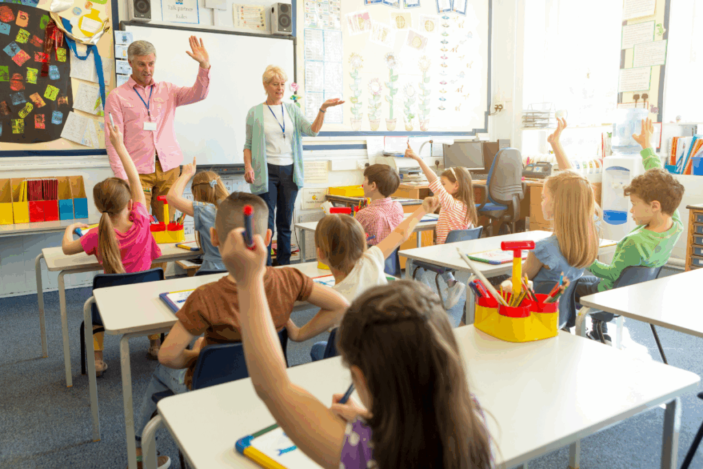 Photo of students raising their hands and looking at two teachers standing at the front of a classroom.